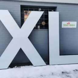 Duże, białe litery XL ustawione pod ścianą budynku z szarej cegły. Na ścianie widoczny szyld firmy Fresh Vision.