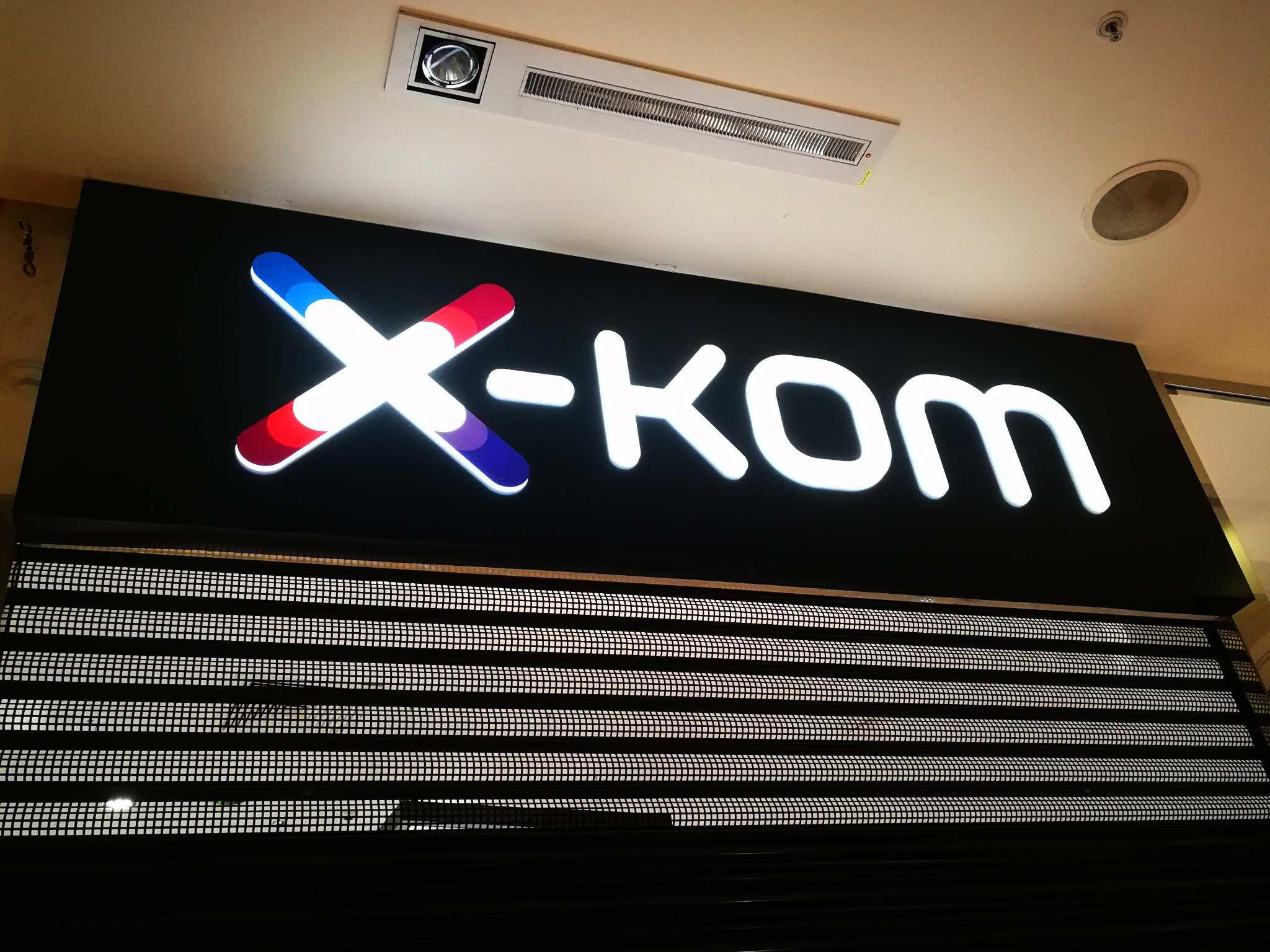 Podświetlany szyld z logo X-KOM w kolorach czerwonym, białym i niebieskim, umieszczony nad metalową żaluzją w sklepie.