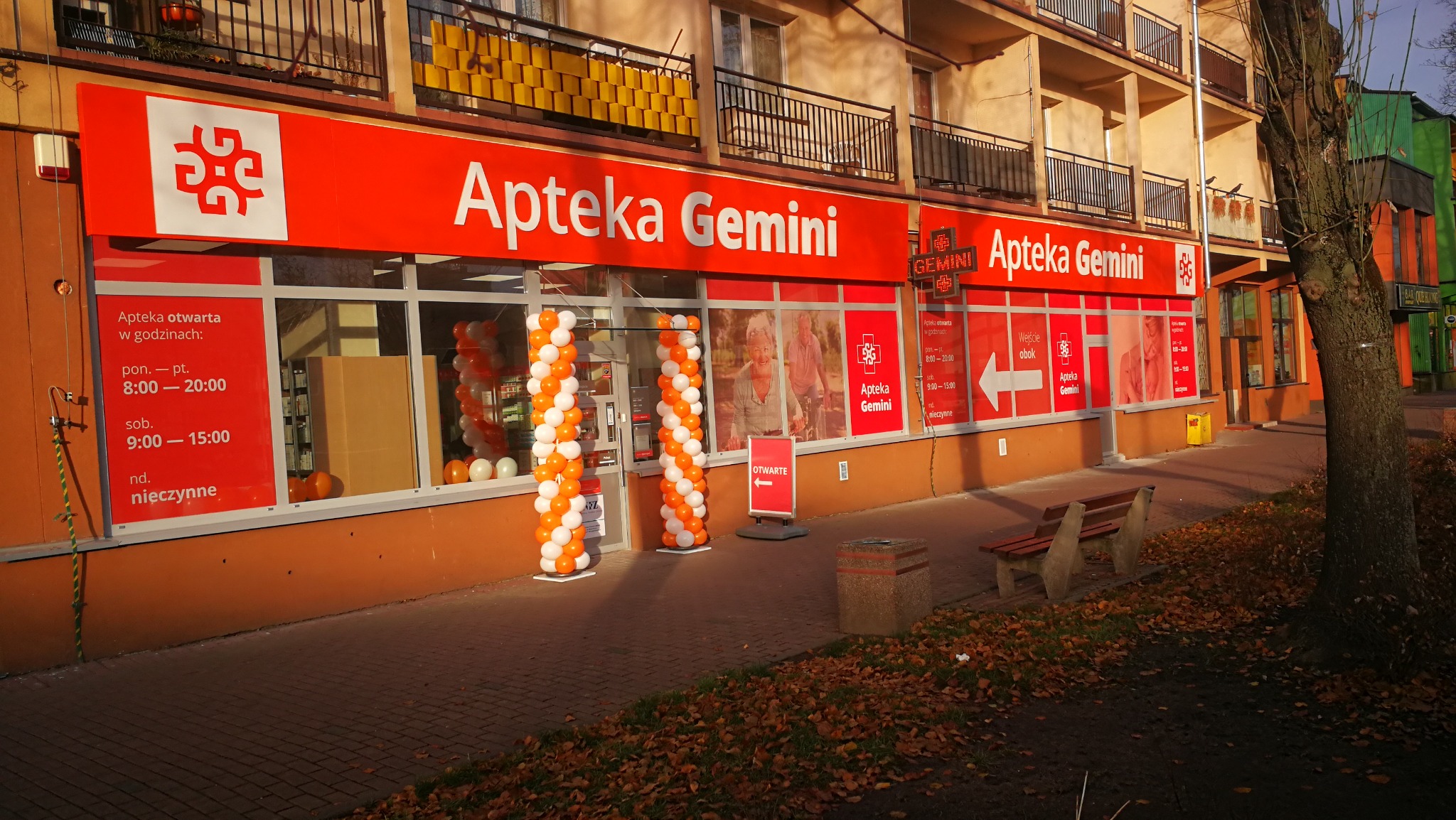 Fasada apteki Gemini z dużym, czerwonym szyldem i logo, okna ozdobione kolumnami z białych i pomarańczowych balonów, widoczne godziny otwarcia na szybach.
