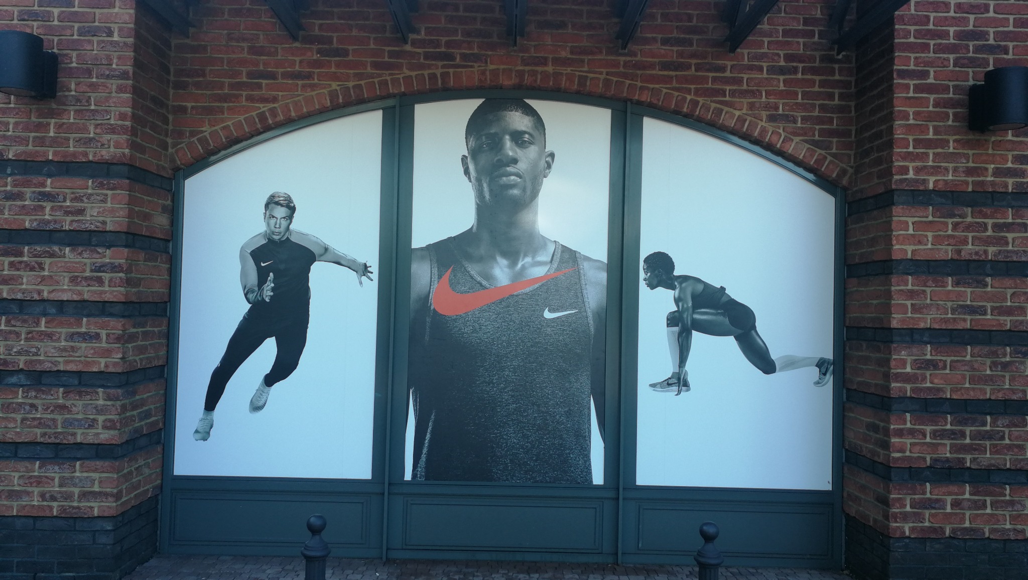 Wielkoformatowy wydruk reklamowy z trzema sportowcami w dynamicznych pozach, umieszczony na fasadzie budynku z cegły.