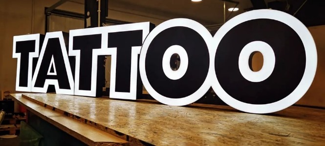 Wykonane z tworzywa, podświetlane litery przestrzenne tworzące napis 'TATTOO' w czarno-białej kolorystyce, stojące na drewnianej powierzchni roboczej.