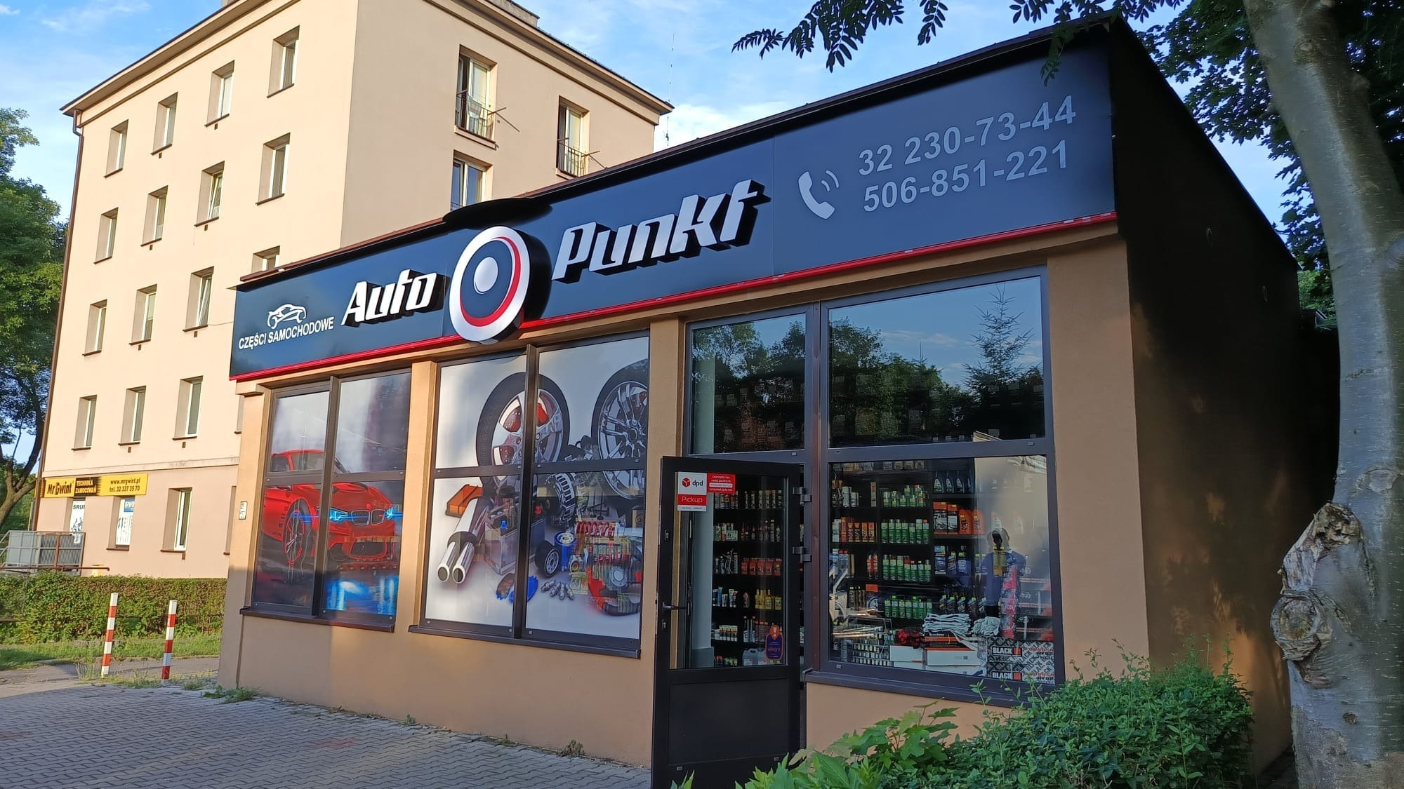Fasada sklepu z częściami samochodowymi 'Auto Punkt' z dużymi witrynami oklejonymi grafikami felg i samochodów, widoczne numery telefonów i logo firmy.