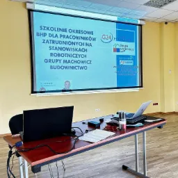 Sala konferencyjna z rzutnikiem wyświetlającym slajd prezentacji o szkoleniu okresowym BHP dla pracowników zatrudnionych na stanowiskach robotniczych Grupy Machowicz Budownictwo, na stole laptop...
