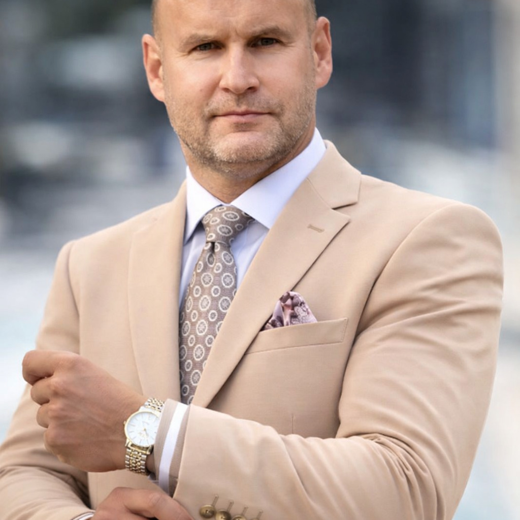 Portret biznesowy mężczyzny w beżowym garniturze, z zegarkiem na ręku, na rozmytym tle. Elegancki krawat i poszetka dodają szyku. Ujęcie od pasa w górę.