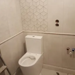 Nowoczesna toaleta kompaktowa w jasnej łazience z częściowo ułożonymi płytkami o wzorze geometrycznym, widoczne narzędzia i drabina.