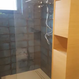 Nowoczesna kabina prysznicowa typu walk-in z odpływem liniowym, obudowana płytkami w odcieniach szarości, z drewnianą szafką łazienkową z boku i poziomnicą na górze.