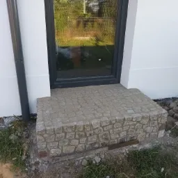 Granitowe schody wejściowe do domu z czarną ramą okienną, widoczny fragment elewacji i rynny, teren w trakcie prac wykończeniowych.