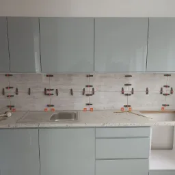 Układanie płytek imitujących drewno na ścianie nad blatem kuchennym, z użyciem pomarańczowych krzyżyków dystansowych, widoczny zlew i szare szafki kuchenne.