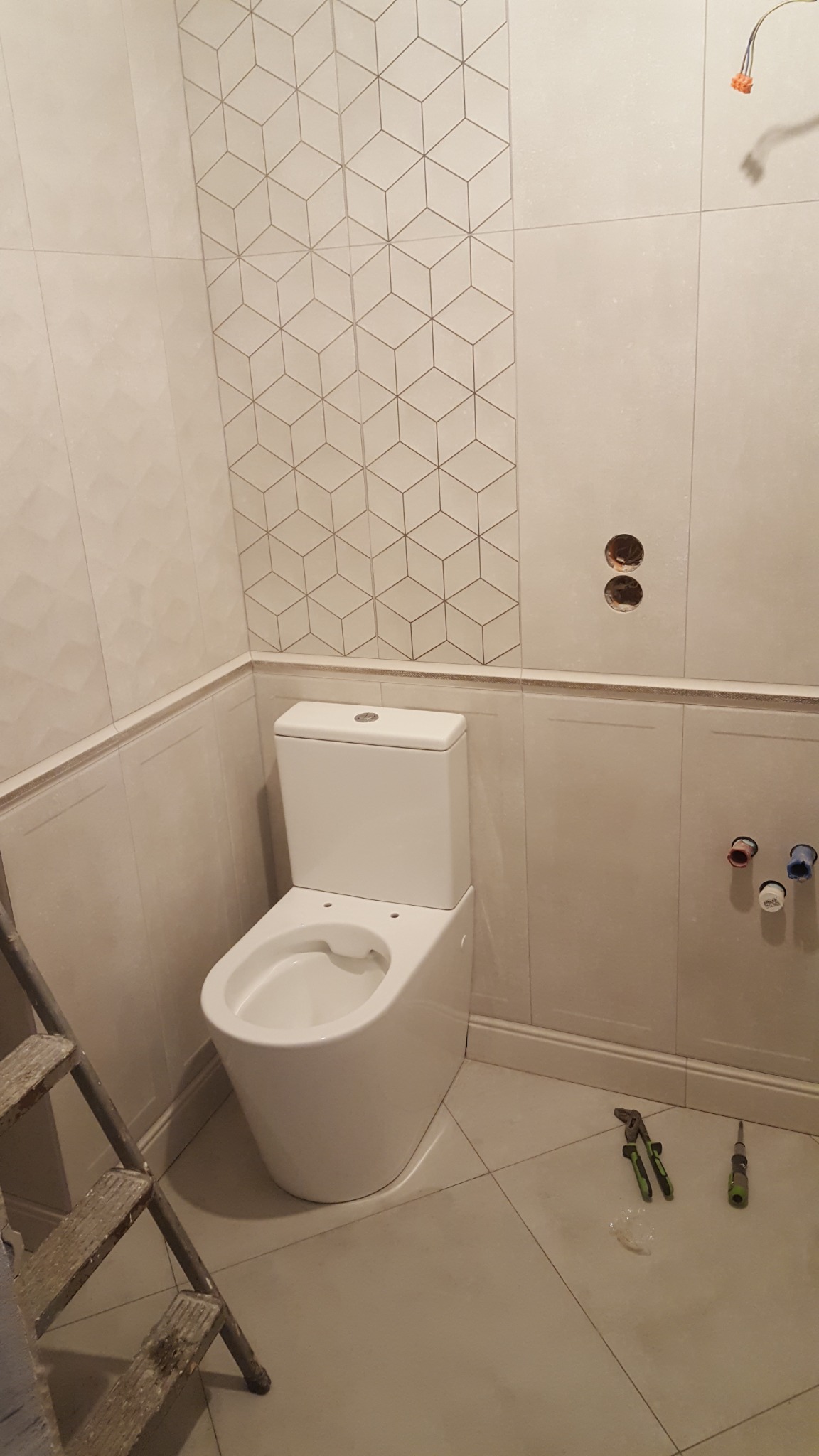 Nowoczesna toaleta kompaktowa w jasnej łazience z częściowo ułożonymi płytkami o wzorze geometrycznym, widoczne narzędzia i drabina.