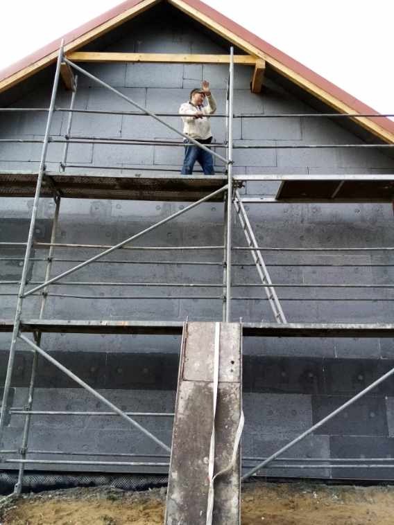 Pracownik na rusztowaniu, ocieplanie ściany zewnętrznej budynku płytami styropianowymi w kolorze grafitowym, widoczny fragment dachu z czerwoną dachówką.