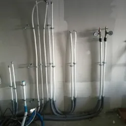 Surowe ściany w trakcie prac instalacyjnych: białe rury przymocowane metalowymi uchwytami, szare izolowane rury łączące, manometry z zaworami na górze, widoczne niedoskonałości ścian i przewody...