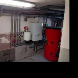 Kocioł gazowy Vaillant z czerwonym zasobnikiem ciepłej wody, widoczne rury instalacji CO oraz wodnej z zaworami termostatycznymi, całość w piwnicy otynkowanej, surowej ścianie.