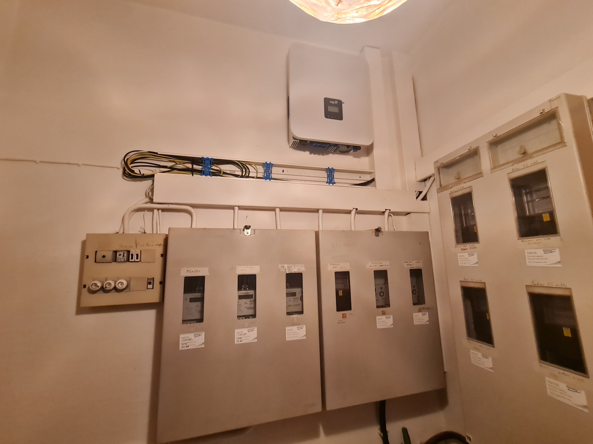 Rozdzielnia elektryczna z bezpiecznikami i liczniki energii elektrycznej w szarej obudowie, zamontowane na ścianie w pomieszczeniu oświetlonym ciepłym światłem.