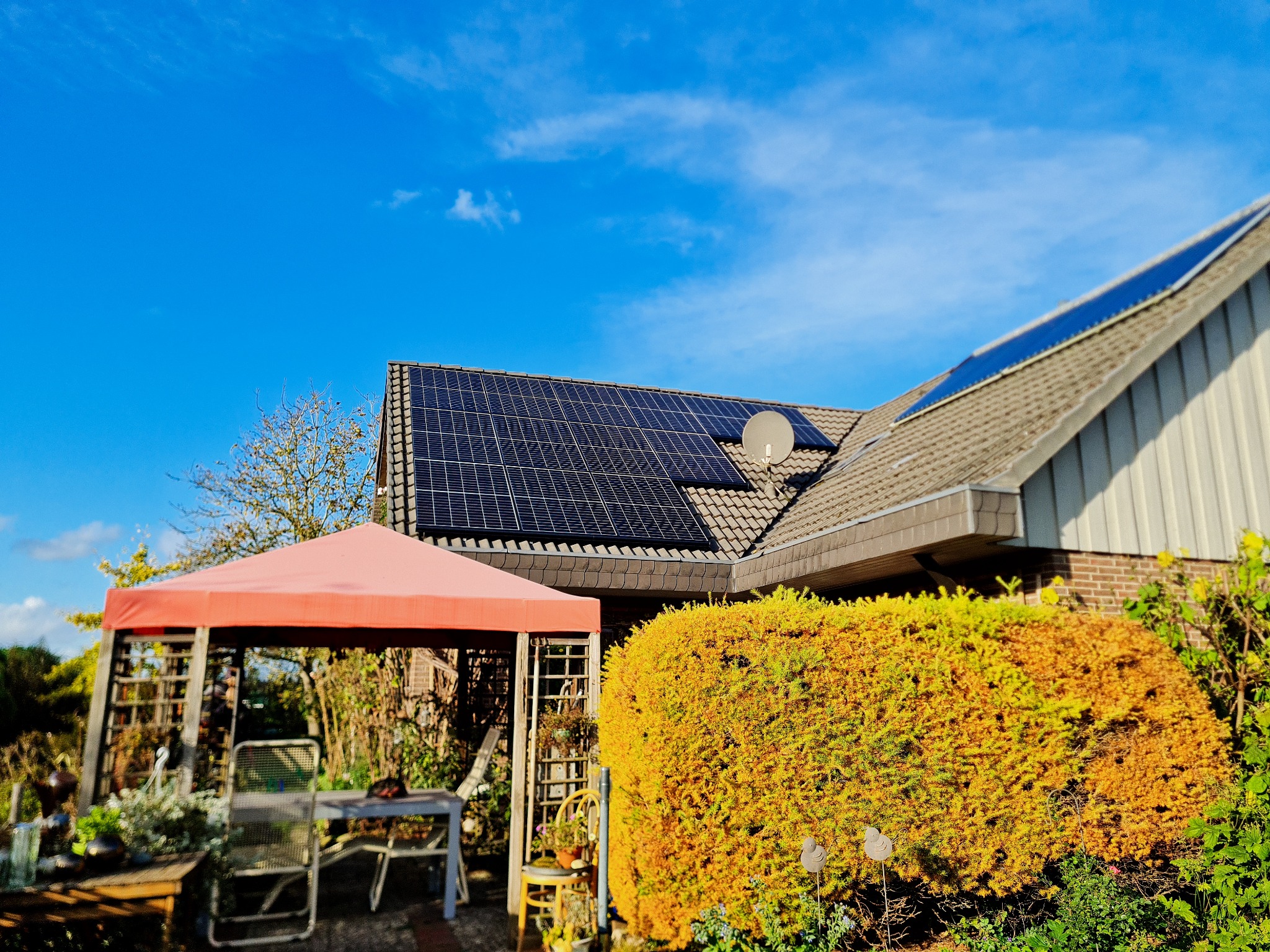 Dom jednorodzinny z panelami fotowoltaicznymi na dachu, antena satelitarna, widok z ogrodu z altaną i bujną roślinnością w słoneczny dzień.