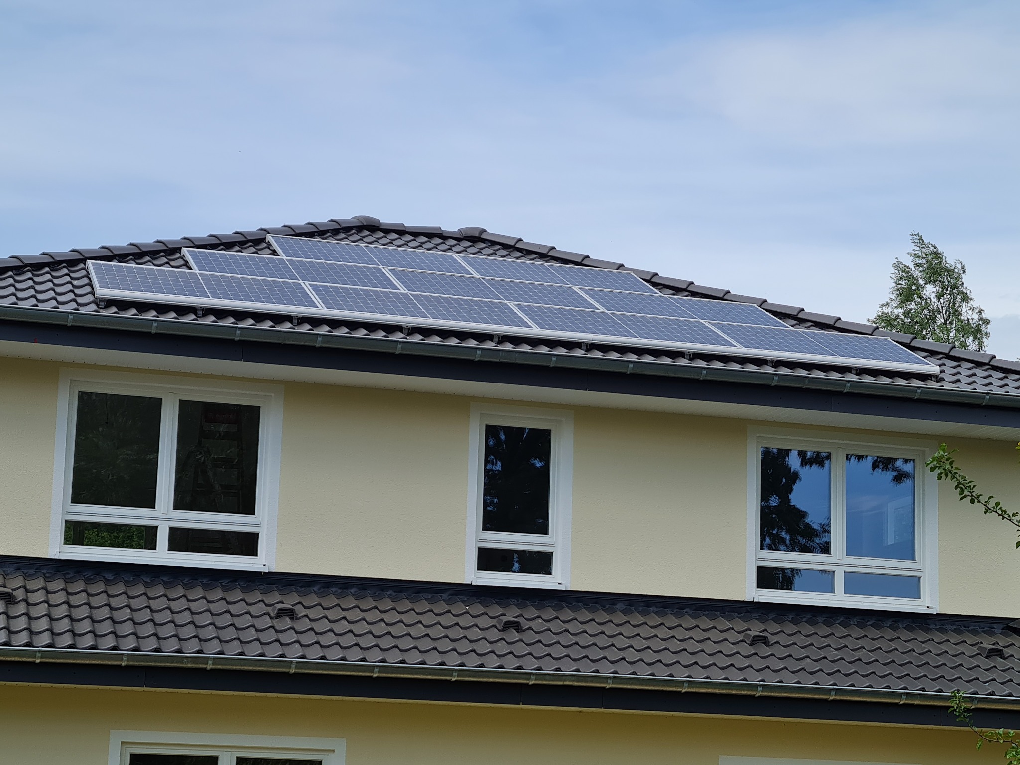 Panele słoneczne zamontowane na dachu dwupiętrowego domu z kremową elewacją i ciemną dachówką, widoczne trzy okna z białymi ramami.