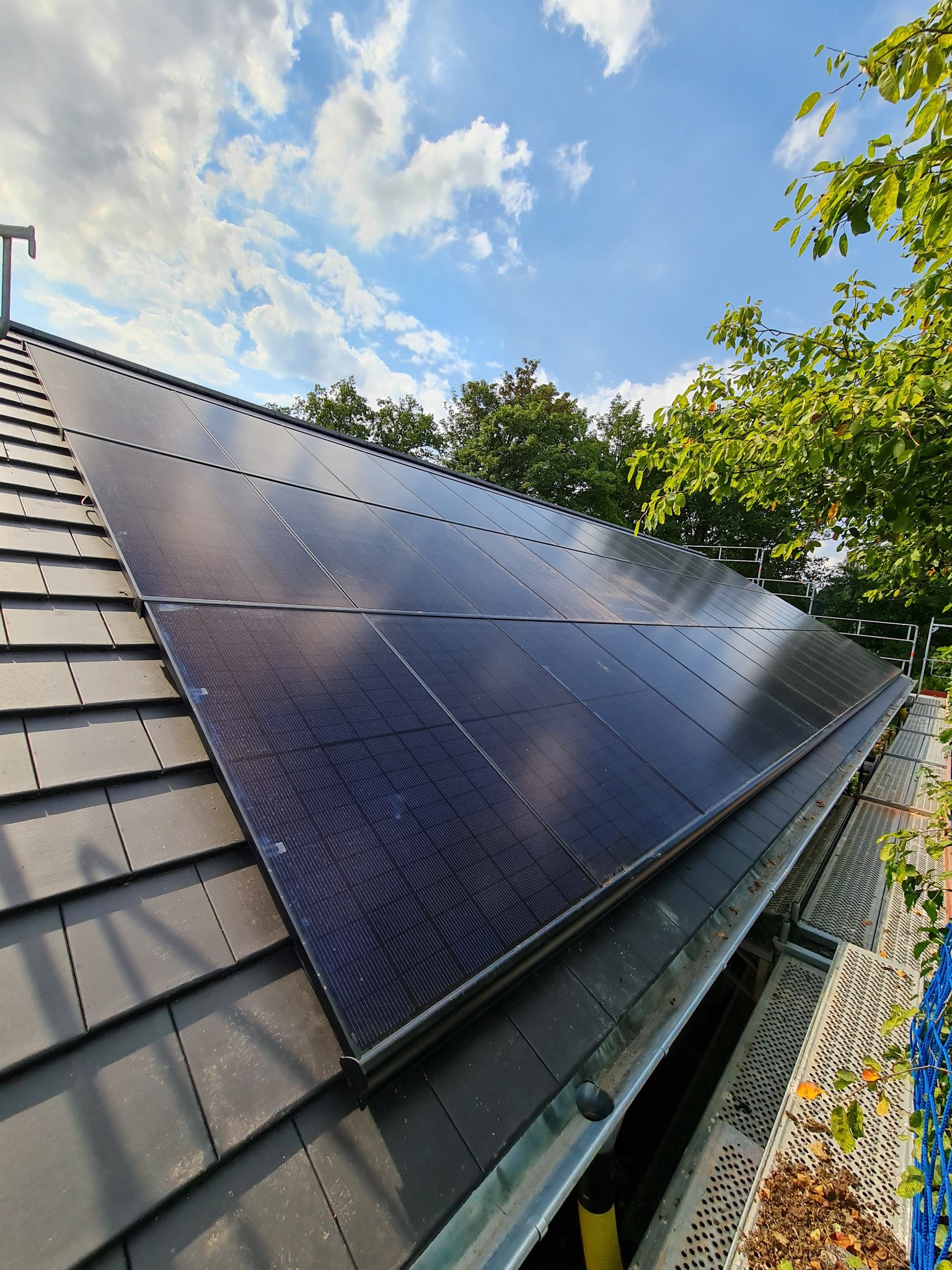 Panel słoneczny zamontowany na dachu pokrytym szarą dachówką, widoczne rusztowanie i fragment nieba z chmurami.