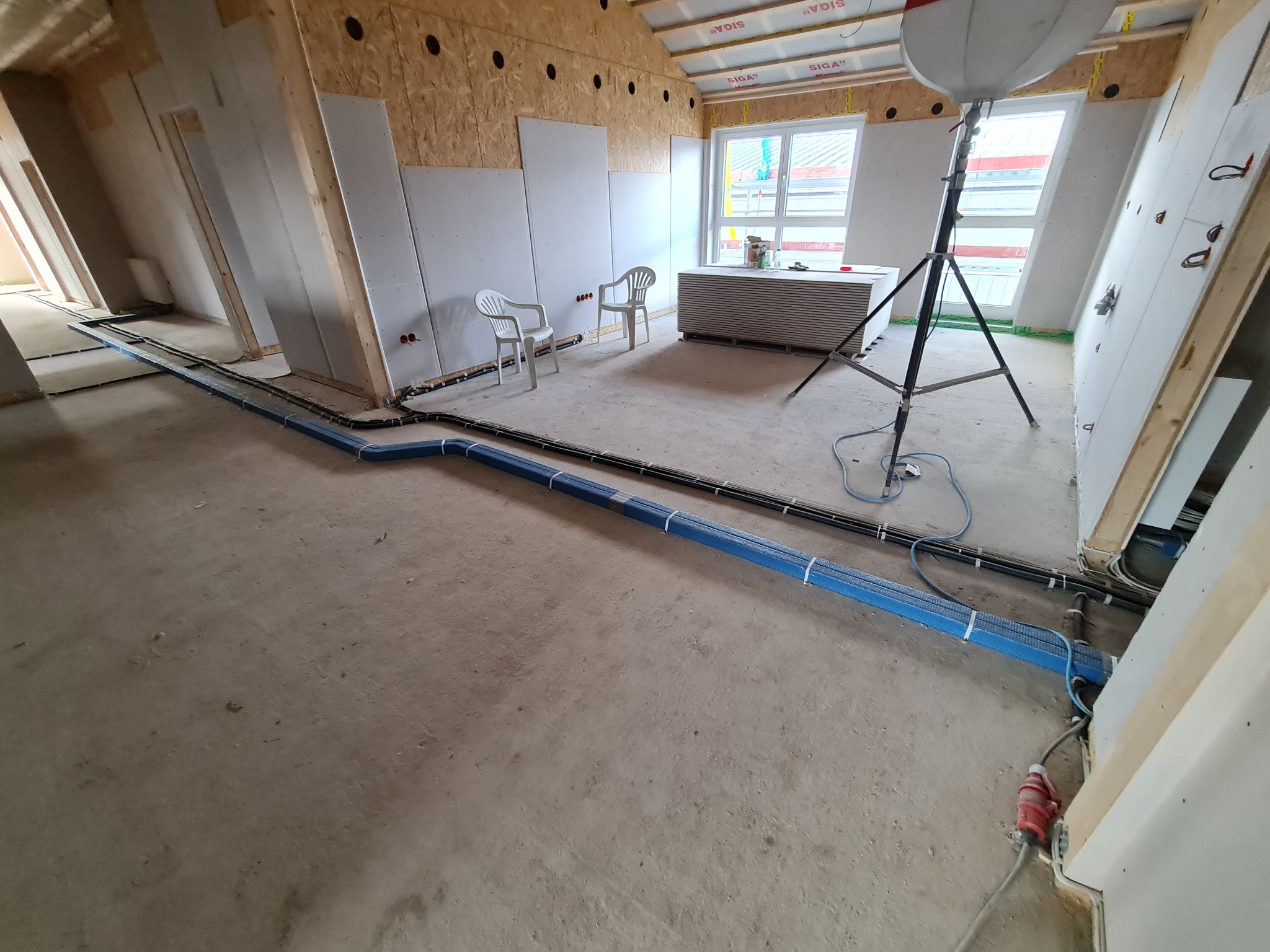 Wnętrze w trakcie remontu w Norderstedt, widoczne prowadzone kable elektryczne w niebieskich korytach podłogowych, płyty gipsowe na ścianach, tymczasowe oświetlenie budowlane i przygotowane płyty...