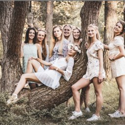 Cezary Gąsiorowski Fotografia - Grupa siedmiu młodych kobiet w letnich sukienkach i wiankach na głowach pozuje w lesie, jedna leży na pniu drzewa.