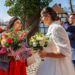 Cezary Gąsiorowski Fotografia - Panna młoda w białej sukni i futrzanym okryciu wymienia uśmiechy z druhną w czerwonej sukience, obie trzymają bukiety kwiatów na tle gości i budynku z muru pruskiego.