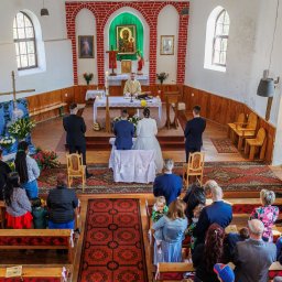 Cezary Gąsiorowski Fotografia - Uroczystość ślubna w kościele, widok z góry na parę młodą klęczącą przed ołtarzem, goście siedzący w ławkach i ksiądz prowadzący ceremonię.
