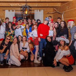 Cezary Gąsiorowski Fotografia - Grupa młodych osób w kostiumach na imprezie w góralskim wnętrzu z drewnianymi ścianami i sufitem, oświetlona wiszącą lampą, jedna osoba trzyma plastikowy topór.