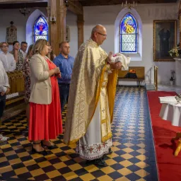 Ksiądz w bogato zdobionej szacie liturgicznej trzyma niemowlę podczas ceremonii w kościele, w tle rodzina i goście, na podłodze czarno-żółta szachownica.