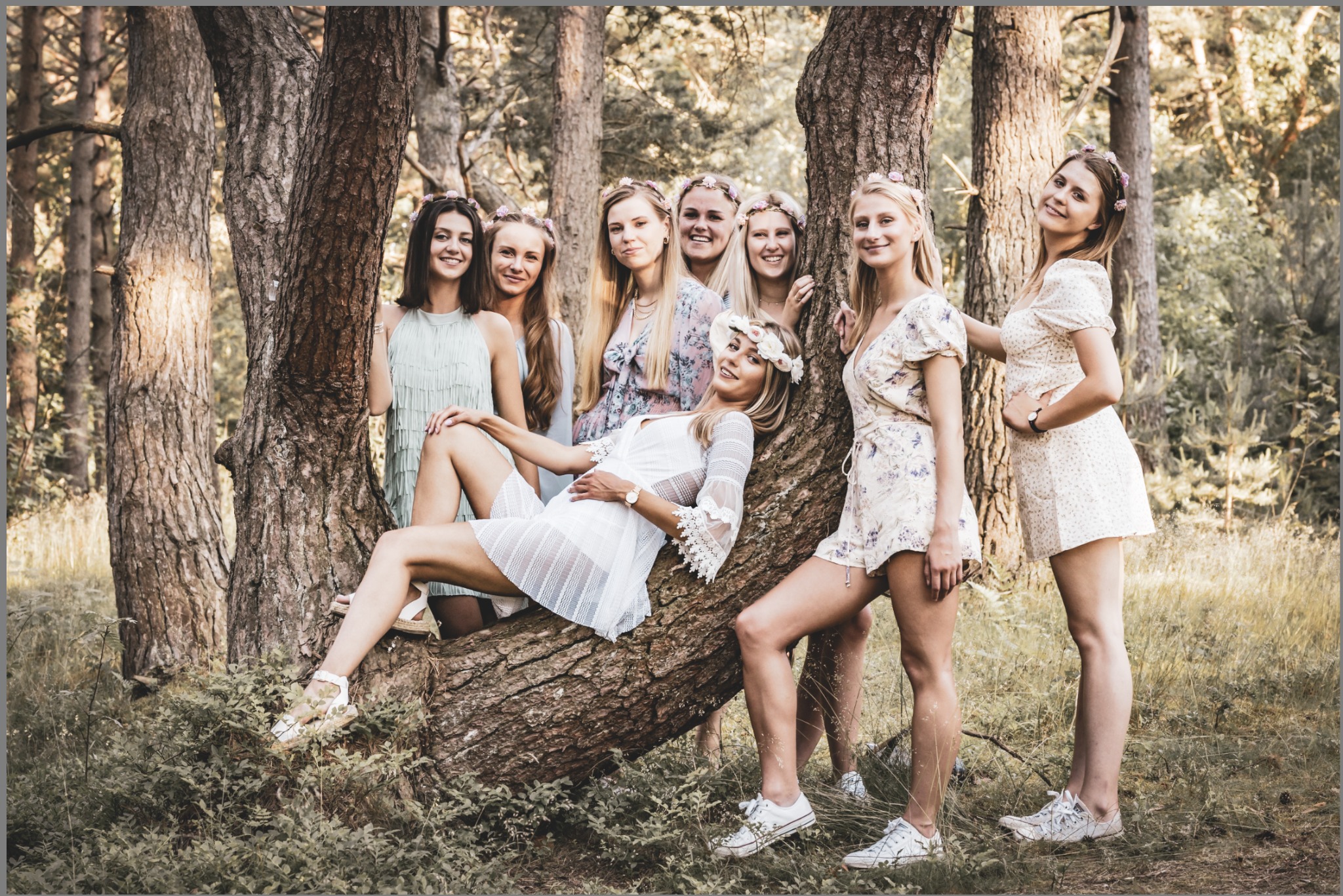 Grupa siedmiu młodych kobiet w letnich sukienkach i wiankach na głowach pozuje w lesie, jedna leży na pniu drzewa.