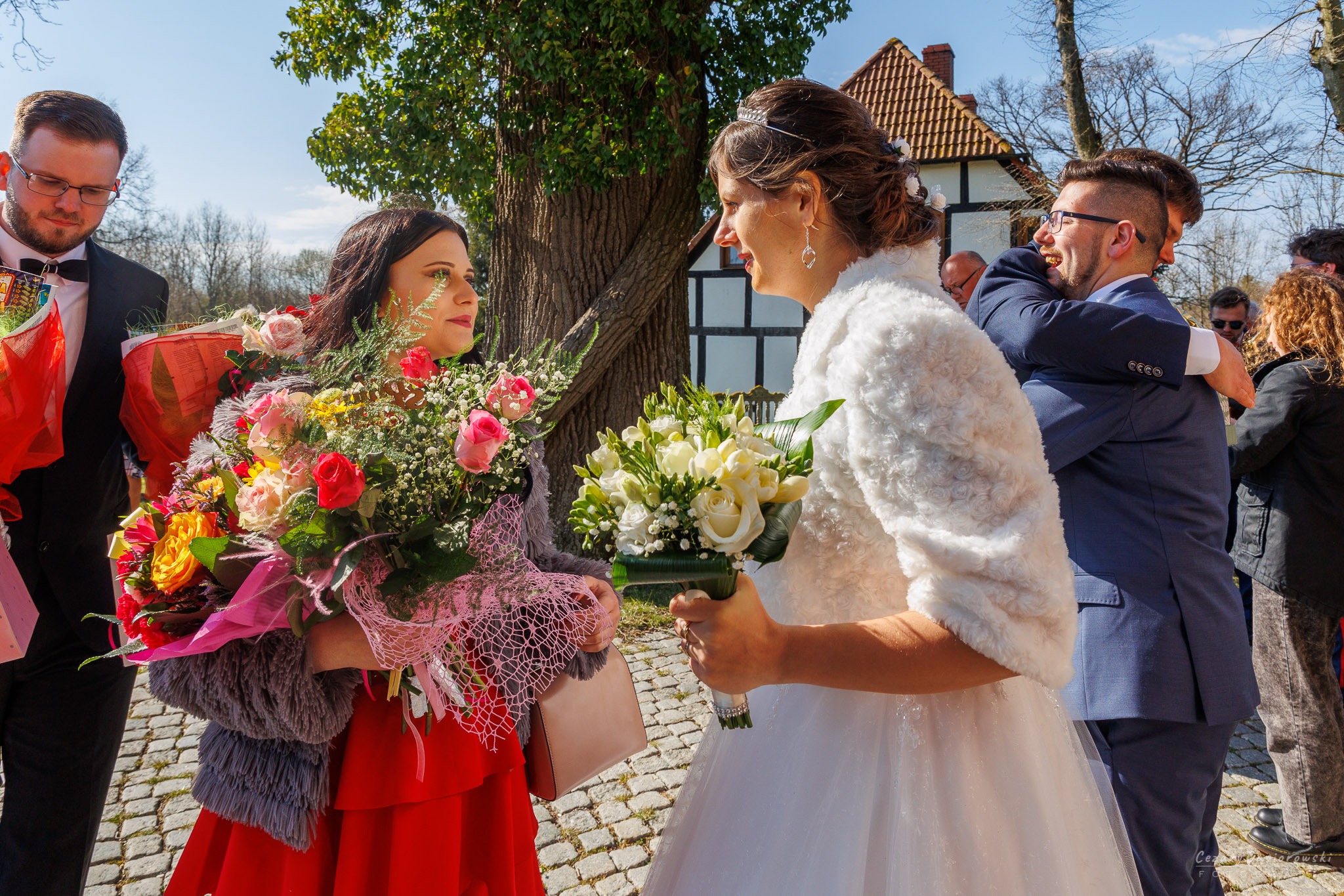Panna młoda w białej sukni i futrzanym okryciu wymienia uśmiechy z druhną w czerwonej sukience, obie trzymają bukiety kwiatów na tle gości i budynku z muru pruskiego.
