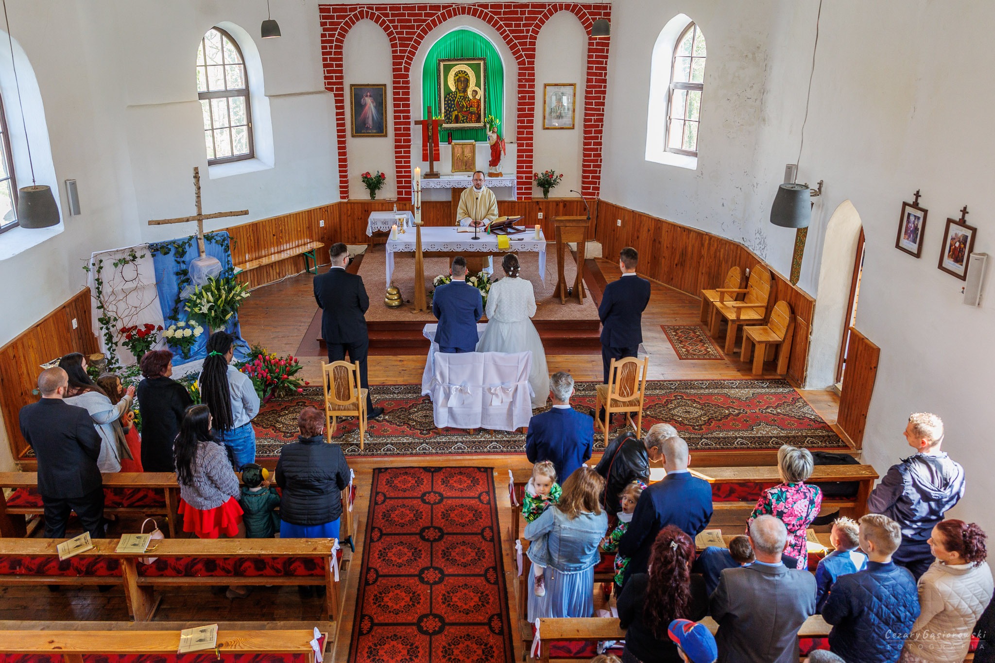 Uroczystość ślubna w kościele, widok z góry na parę młodą klęczącą przed ołtarzem, goście siedzący w ławkach i ksiądz prowadzący ceremonię.