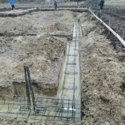 Zalane wykopane rowy zbrojone stalą na fundamenty domu jednorodzinnego, widoczni pracownicy budowlani w oddali.