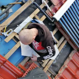 Prace na dachu: montaż izolacji pod panelami solarnymi, widok z góry na dekarza w trakcie pracy, elementy konstrukcji dachu, czerwona dachówka i narzędzia.