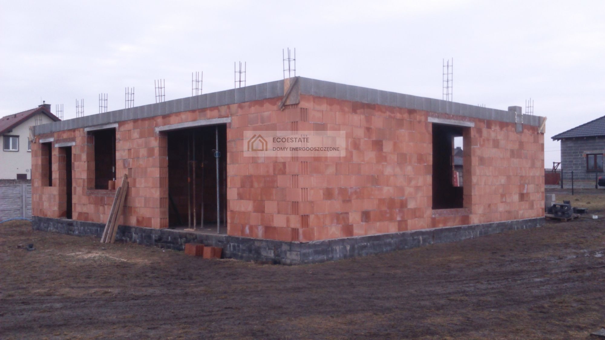Surowy stan domu z czerwonej cegły z widocznym logo ECOESTATE na ścianie, z otworami okiennymi i zbrojeniami na dachu, w tle inne domy.