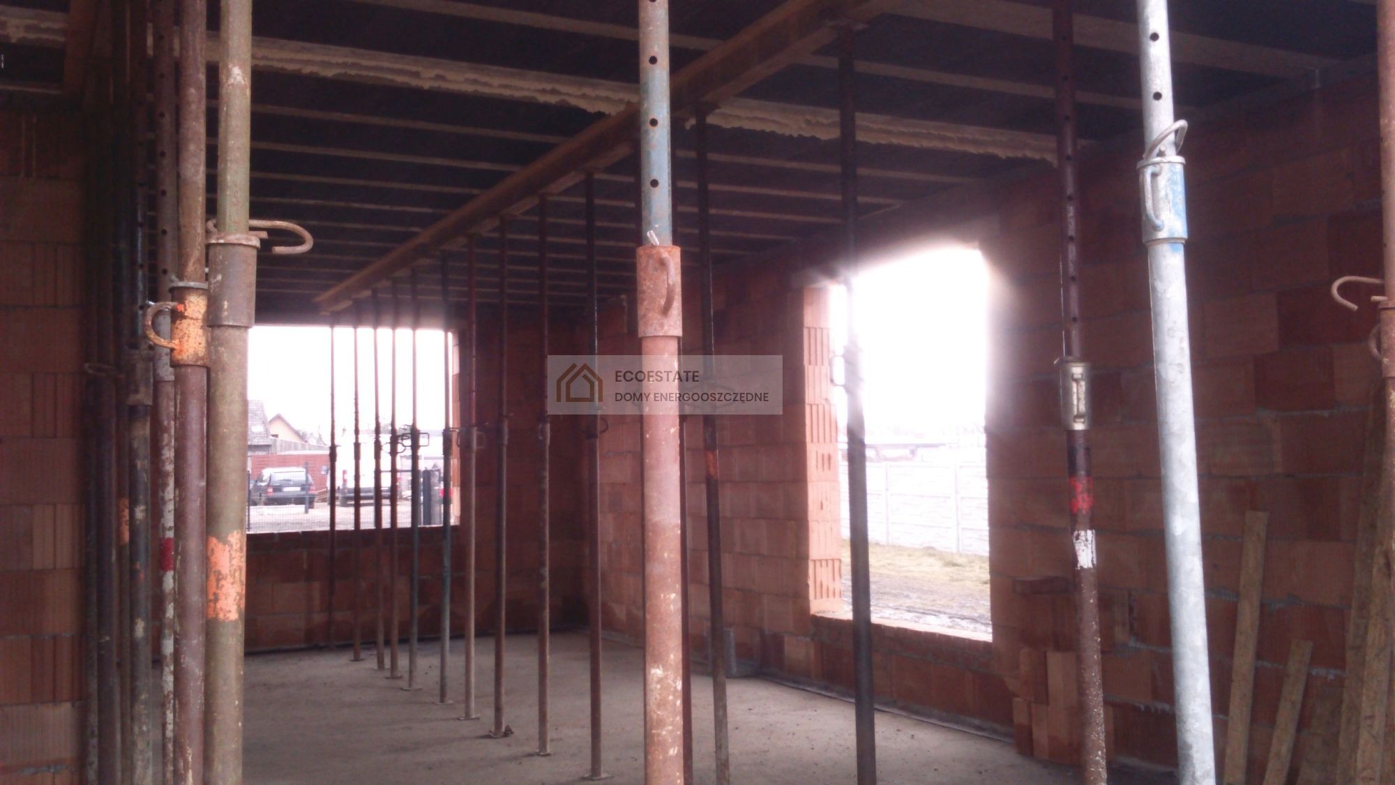 Wnętrze w trakcie budowy z cegły ceramicznej, z widocznymi podporami stropowymi i oknami bez ram, w tle krajobraz wiejski.
