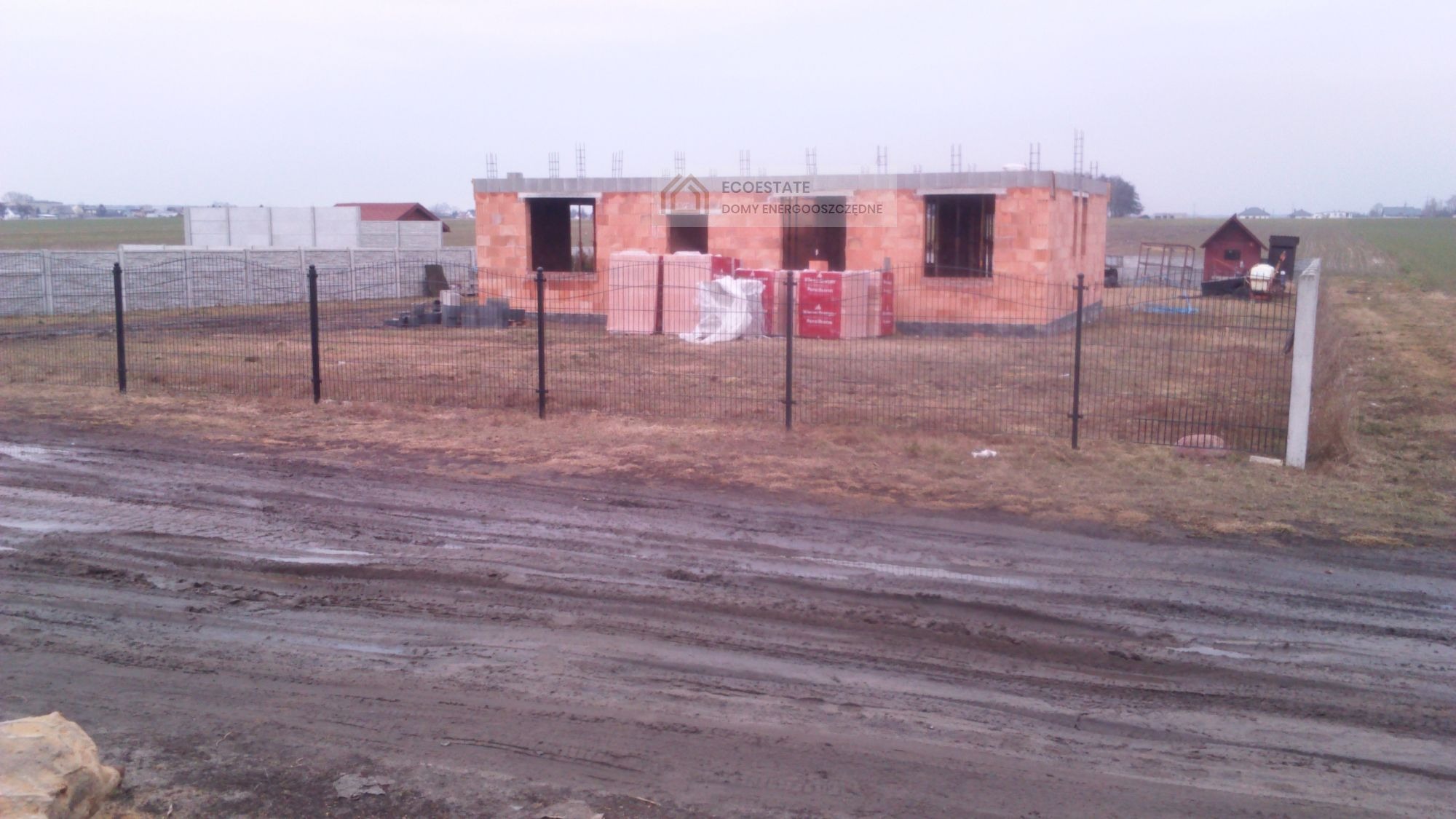 Budowa domu z czerwonej cegły, widoczne materiały budowlane, ogrodzenie z siatki i błotnista droga dojazdowa w pochmurny dzień.