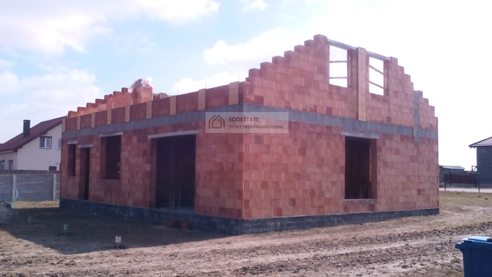 Surowy stan domu z czerwonej cegły z widocznym logo 'ECOESTATE DOMY ENERGOOSZCZĘDNE' na ścianie, ukazujący etap budowy z niezadaszonym dachem i otworami okiennymi.