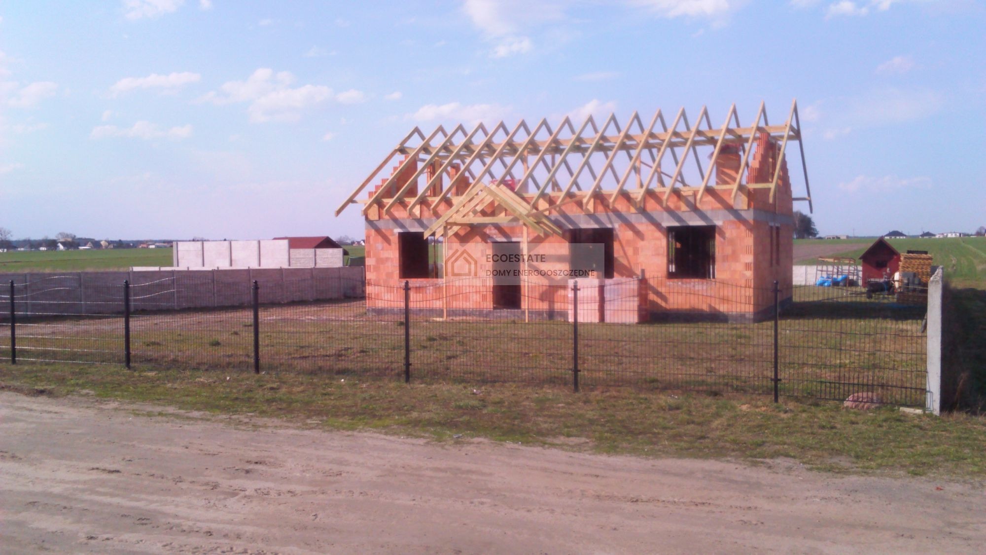 Niedokończony budynek z czerwonej cegły z drewnianą konstrukcją dachu, ogrodzony czarnym metalowym płotem, na tle krajobrazu z polami i innymi zabudowaniami.