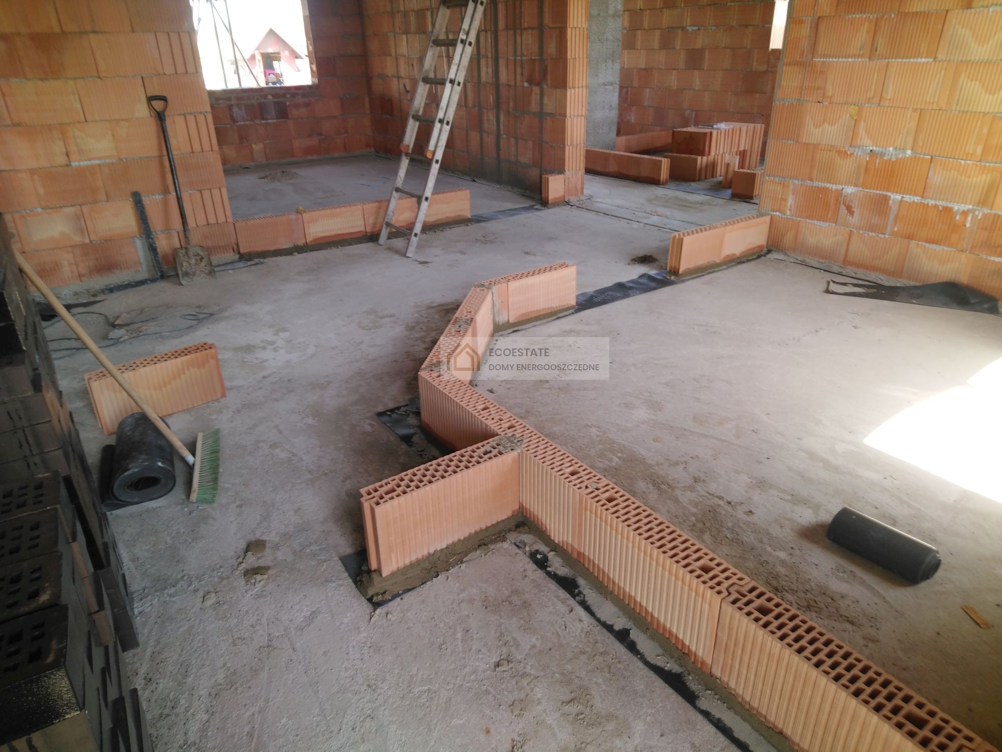 Wnętrze w trakcie budowy z cegły ceramicznej: wznoszone ściany, widoczny fundament z izolacją, drabina oparta o ścianę, narzędzia budowlane i cegły na podłodze.