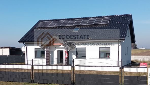 Nowoczesny dom murowany z panelami słonecznymi na dachu, widok z zewnątrz przez metalowe ogrodzenie.