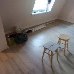 Poddasze po remoncie z nową podłogą z paneli, widoczne dwa taborety, narzędzia i listwy przypodłogowe przygotowane do montażu.