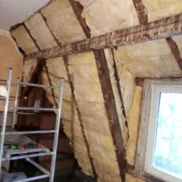 Poddasze w trakcie ocieplania wełną mineralną, widoczne drewniane belki konstrukcyjne, drabina aluminiowa z narzędziami i okno