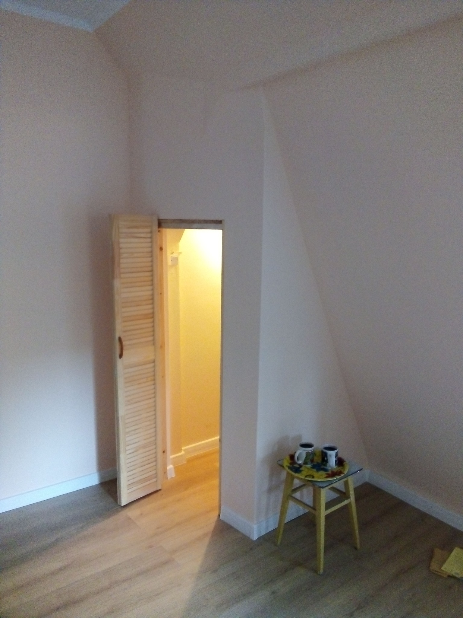 Poddasze po remoncie z drewnianą podłogą, otwartymi lamelkowymi drzwiami do garderoby i małym stolikiem z dwoma kubkami.