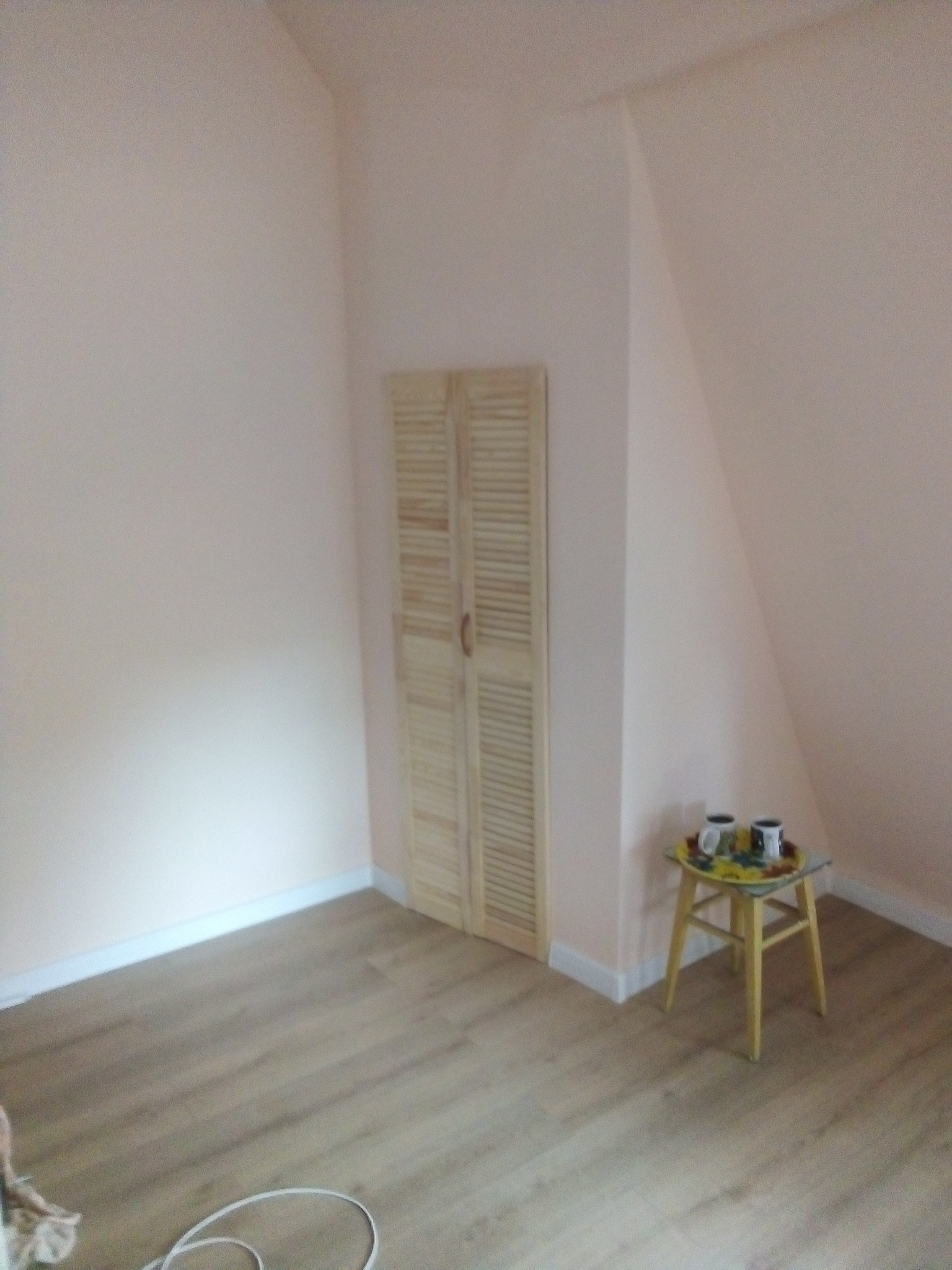 Poddasze z jasnoróżowymi ścianami, drewnianą podłogą i szafką z żaluzjowymi drzwiami, na żółtym stołku taca z dwoma kubkami.