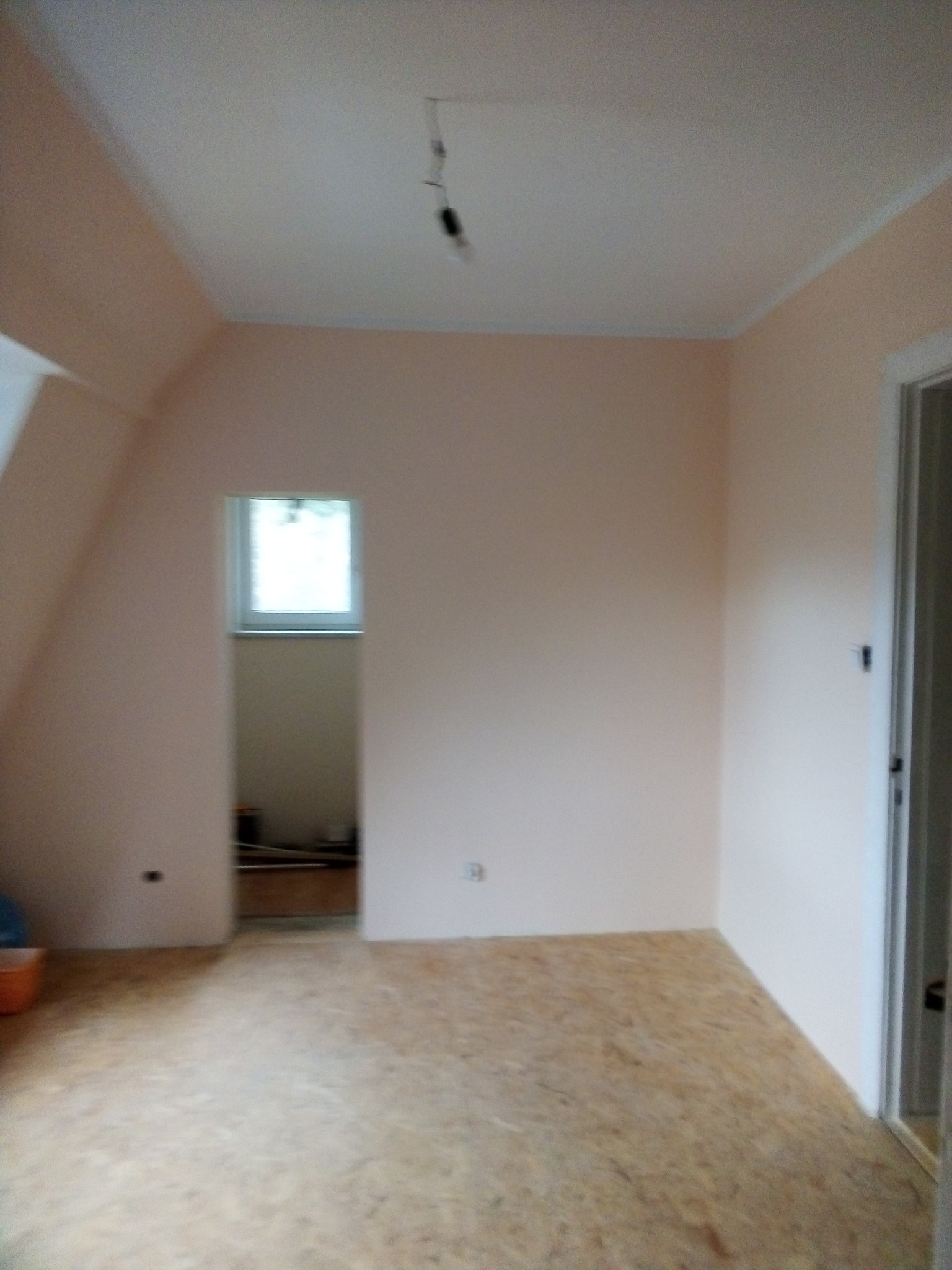 Poddasze po szpachlowaniu i malowaniu na jasny róż, z oknem i podłogą z płyt OSB, przygotowane do dalszych prac wykończeniowych, widoczna surowa żarówka.