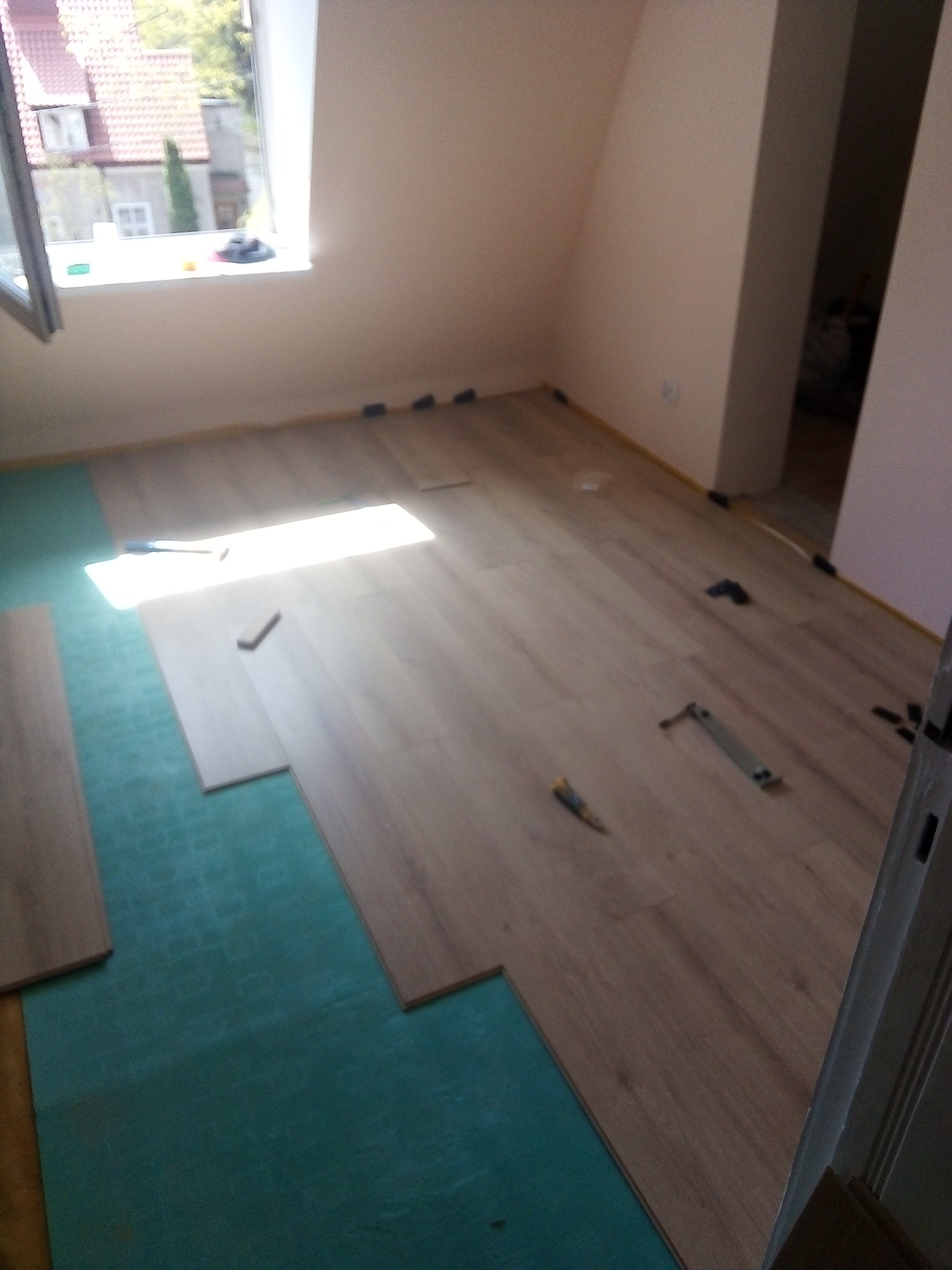 Układanie paneli podłogowych w pokoju na zielonym podkładzie, widoczne narzędzia i elementy mocujące, w tle okno i fragment budynku.