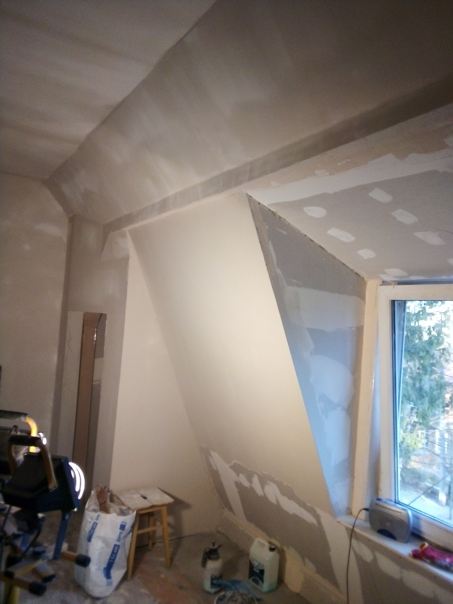 Poddasze w trakcie prac wykończeniowych: ściany i sufit częściowo pokryte szpachlą, widoczne narzędzia, worek gipsu, okno z widokiem na drzewa.