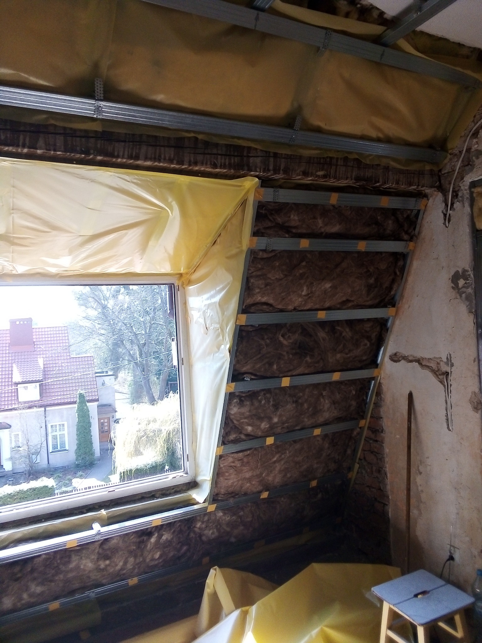 Poddasze w trakcie ocieplania wełną mineralną, widoczne metalowe profile, folia ochronna na oknie oraz fragment starej ściany z ubytkami tynku.