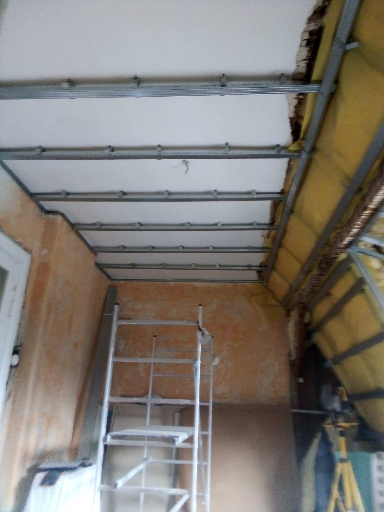 Poddasze w trakcie prac: widoczna konstrukcja metalowa sufitu podwieszanego z częściowo zamontowanymi płytami gipsowymi, ocieplenie wełną mineralną na skosach dachu oraz drabina aluminiowa.