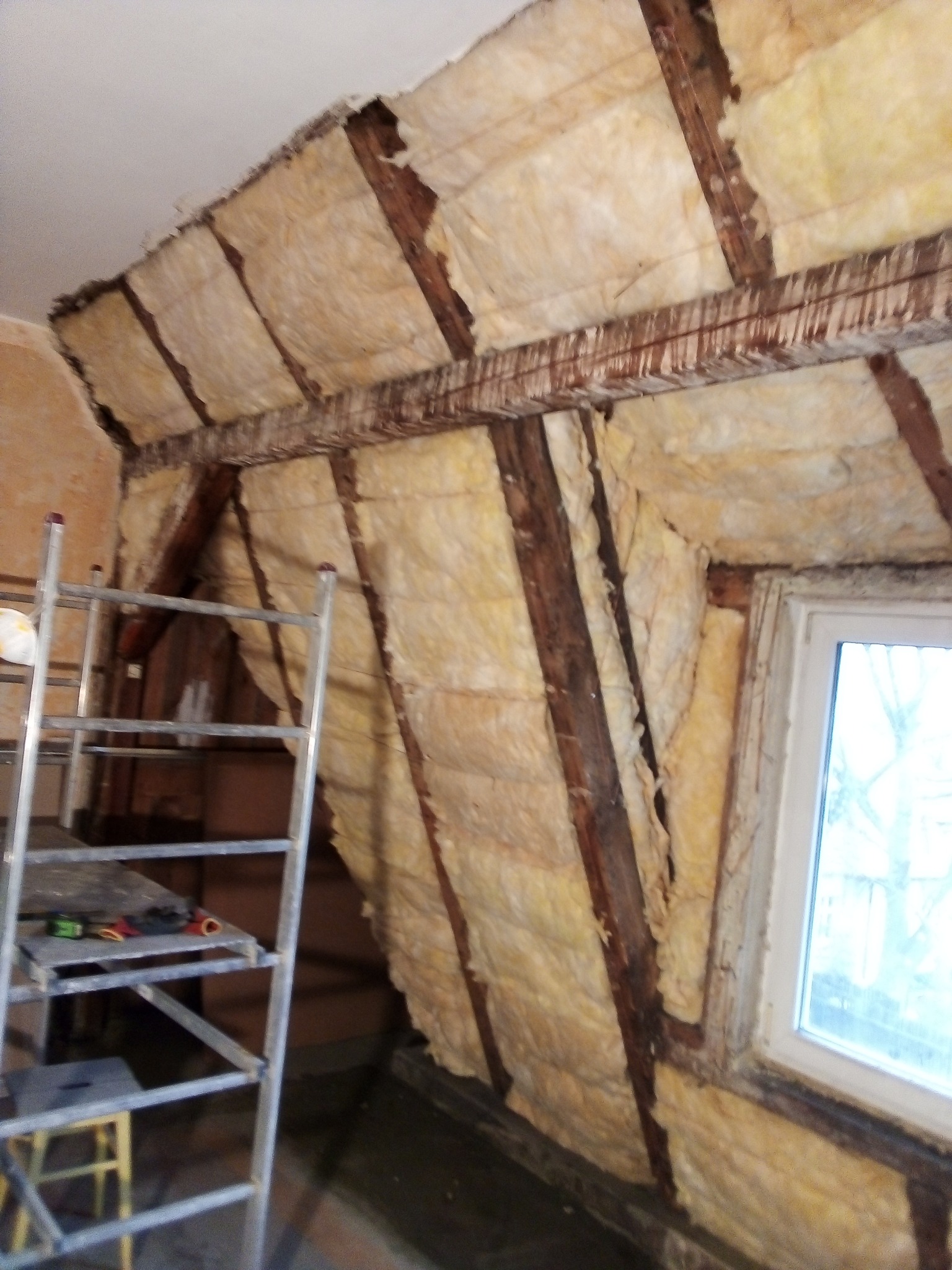 Poddasze w trakcie ocieplania wełną mineralną, widoczne drewniane belki konstrukcyjne, drabina aluminiowa z narzędziami i okno
