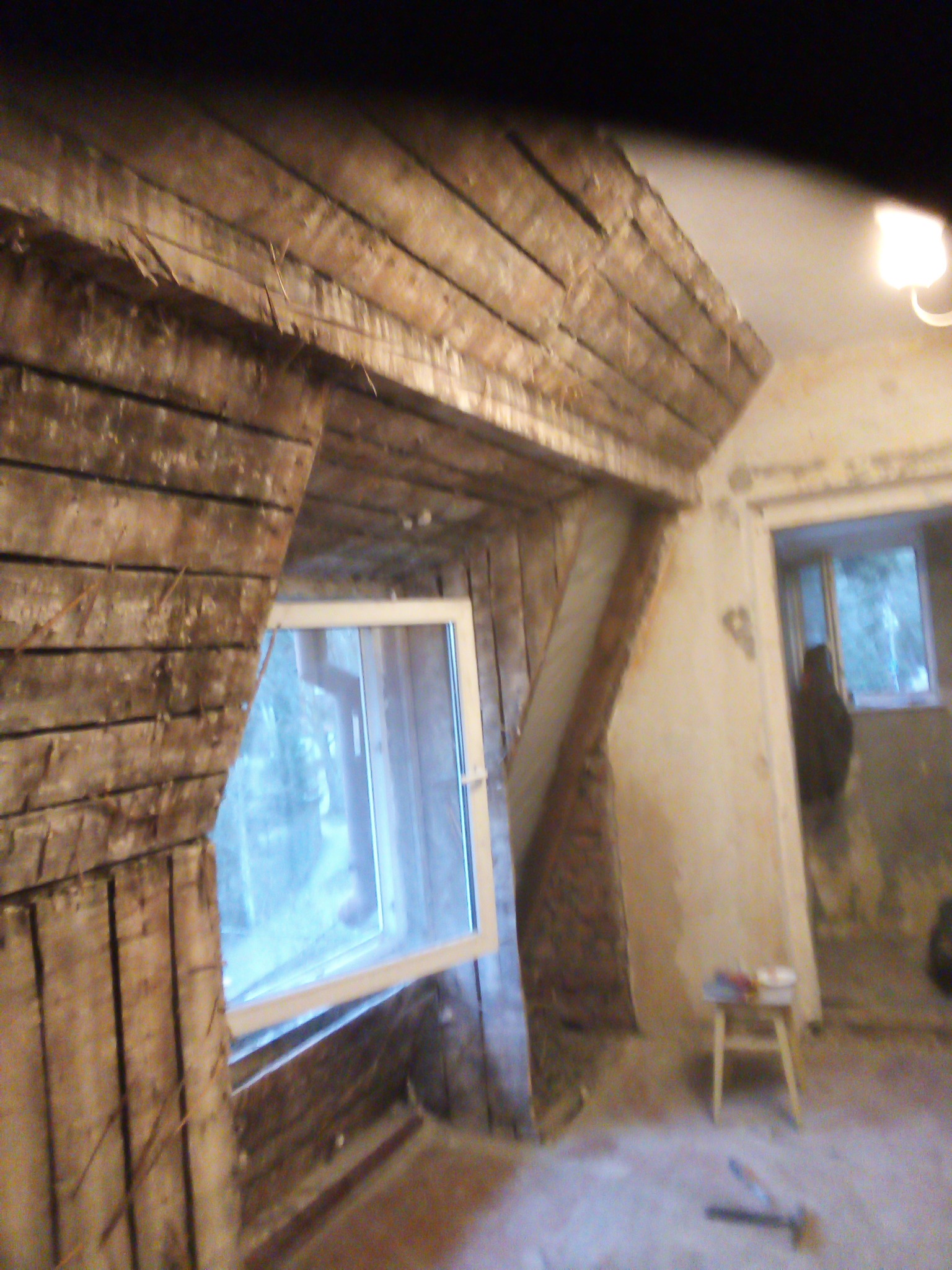 Poddasze w trakcie remontu z widoczną drewnianą konstrukcją ściany szczytowej i nowym oknem dachowym, na podłodze leżą narzędzia, a w tle widać fragment pomieszczenia z krzesłem i osobą w oknie.