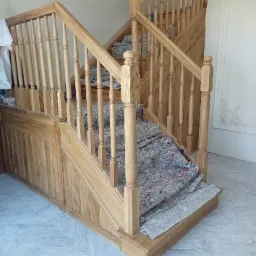 Drewniane schody z balustradą w trakcie montażu, zabezpieczone folią malarską, widoczne zdobienia na ścianie i żyrandol kryształowy.